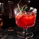 A Pomegranate Boulevardier cocktail in a cut crystal glass, garnished with a smoking sage sprig and ice, with dried petals and a pitcher in a dark, moody setting.