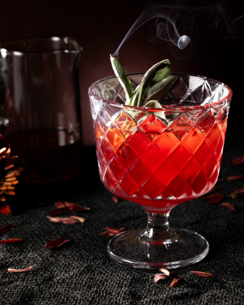 A Pomegranate Boulevardier cocktail in a cut crystal glass, garnished with a smoking sage sprig and ice, with dried petals and a pitcher in a dark, moody setting.