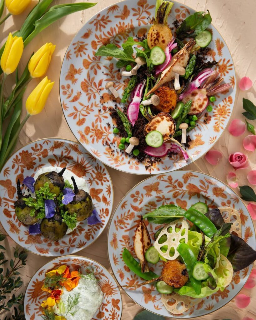 A collection of three plates on a table with a floral pattern and yellow tulips, featuring various roasted and fresh root vegetables with herbs, dressings, and garnishes.