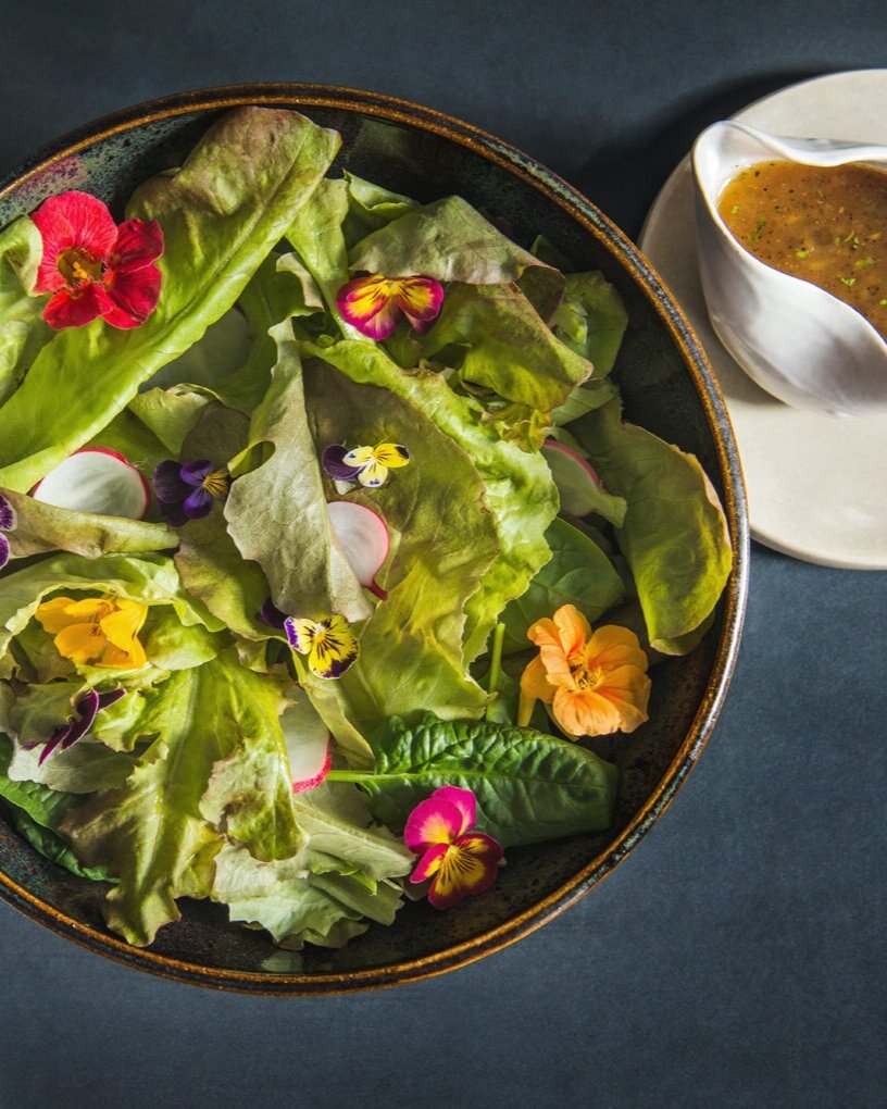 A salad with lettuce, radishes, edible flowers, and an apple bourbon dressing all in a black bowl on a dark blue table.