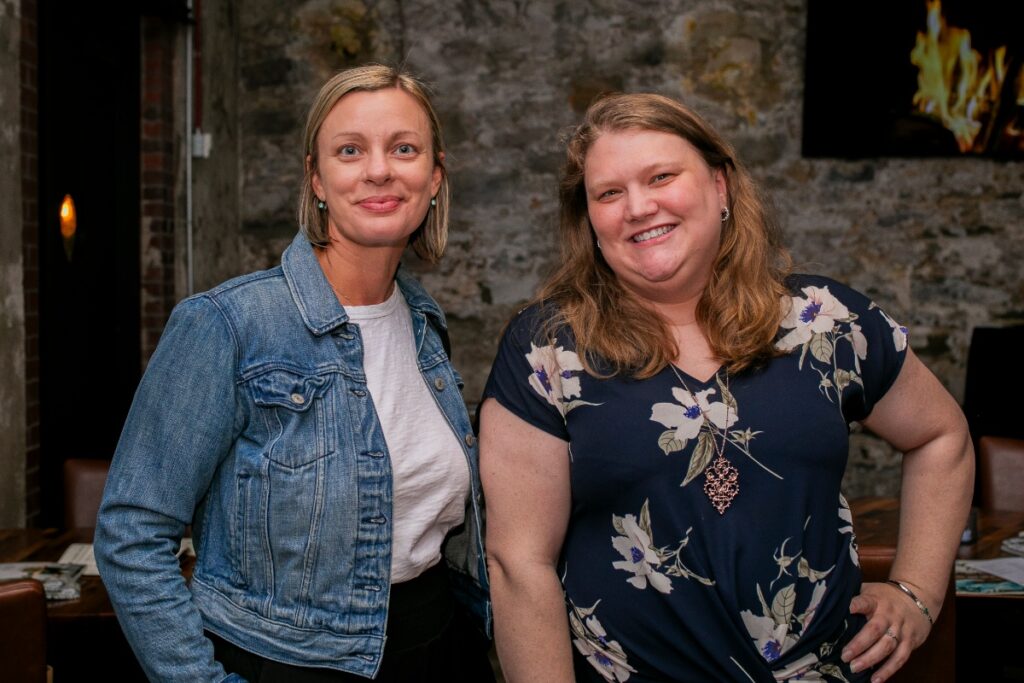 Two smiling women, Sarah Mayer in a denim jacket and Kristen Nuss in a floral top, standing together at a non-profit networking mixer.