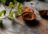 How to Bake with Climate Change in Mind A dark, moody overhead shot of a gray and blue tiled surface dusted with cocoa powder, featuring a small bowl of cocoa powder and a vine with green leaves in the upper corner, suggesting baking.