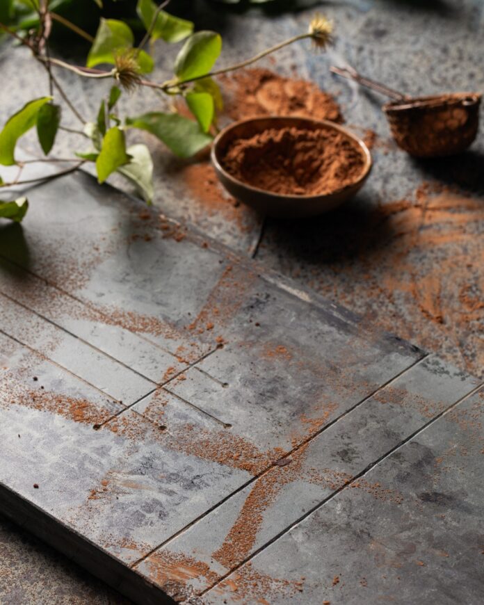 Climate Change Baking A dark, moody overhead shot of a gray and blue tiled surface dusted with cocoa powder, featuring a small bowl of cocoa powder and a vine with green leaves in the upper corner, suggesting baking.