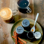 Three small mason jars filled with coffee syrups sit on a plate in front of two cups of coffee.