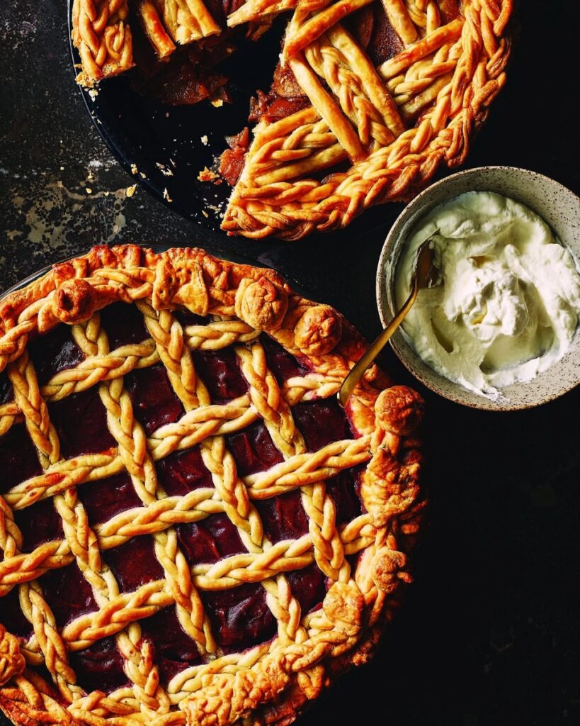 Two gourmet fruit pies by Selina Progar, featuring intricate braided lattice crusts and rich fillings, one whole and one with a slice missing, served next to a bowl of whipped cream.