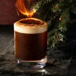 A Smoked Vanilla Carajillo cocktail in a rocks glass with a layer of foam and a burning, cinnamon-dusted orange peel garnish, set next to a pine garland.