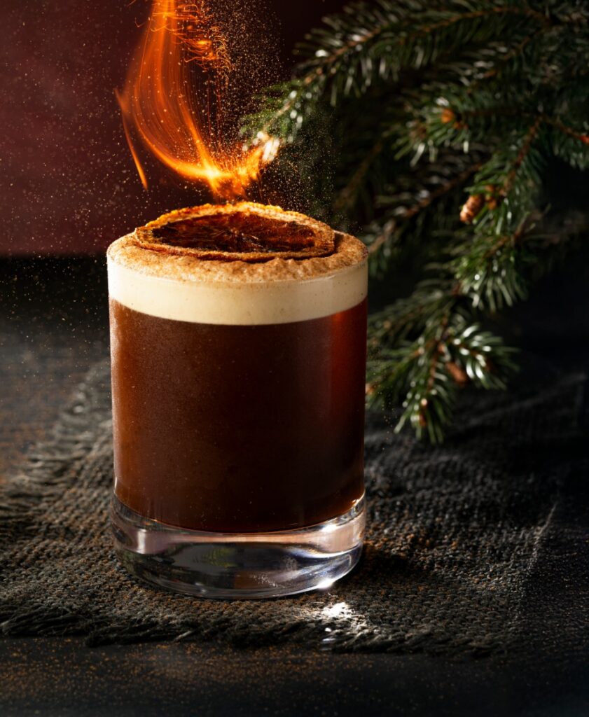 A Smoked Vanilla Carajillo cocktail in a rocks glass with a layer of foam and a burning, cinnamon-dusted orange peel garnish, set next to a pine garland.
