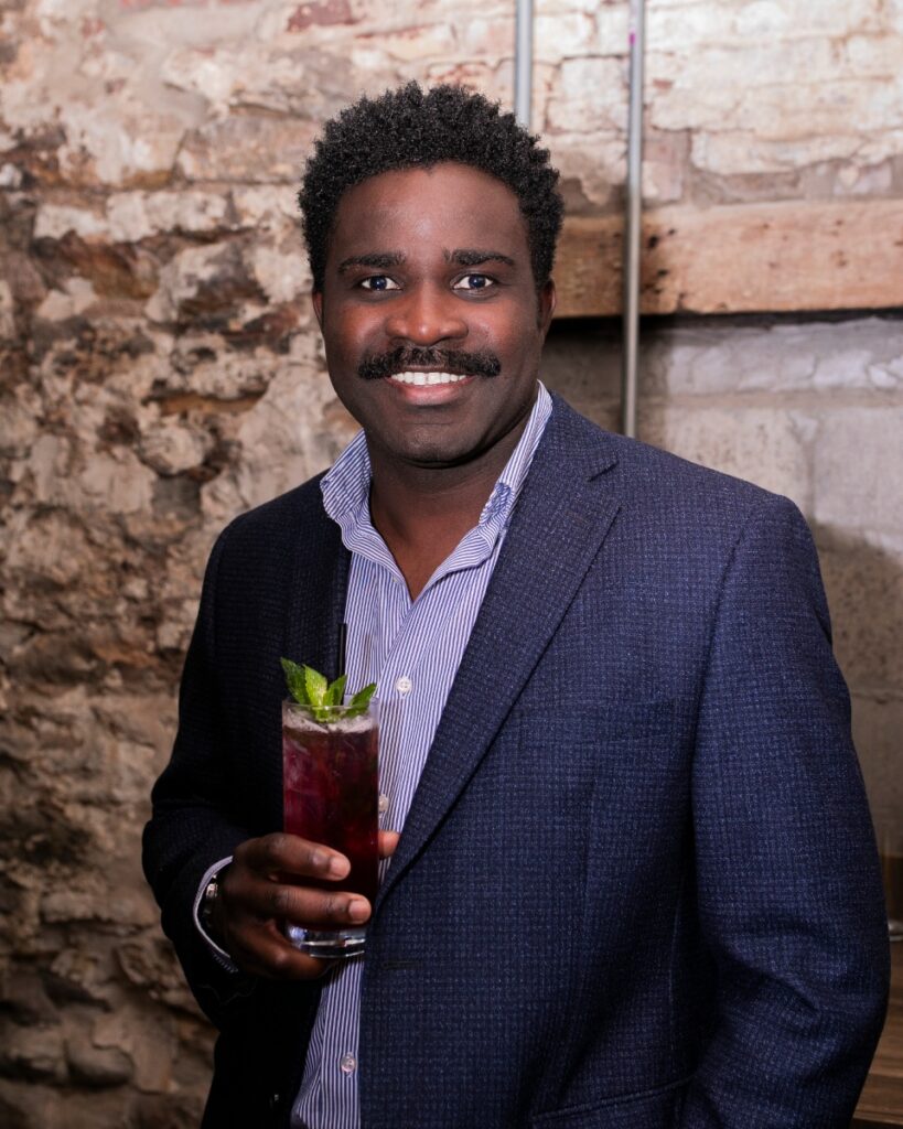 Sony Ton-Aime, a Black man with a mustache, wearing a blue blazer and striped shirt while holding a cocktail, at a non-profit networking event.