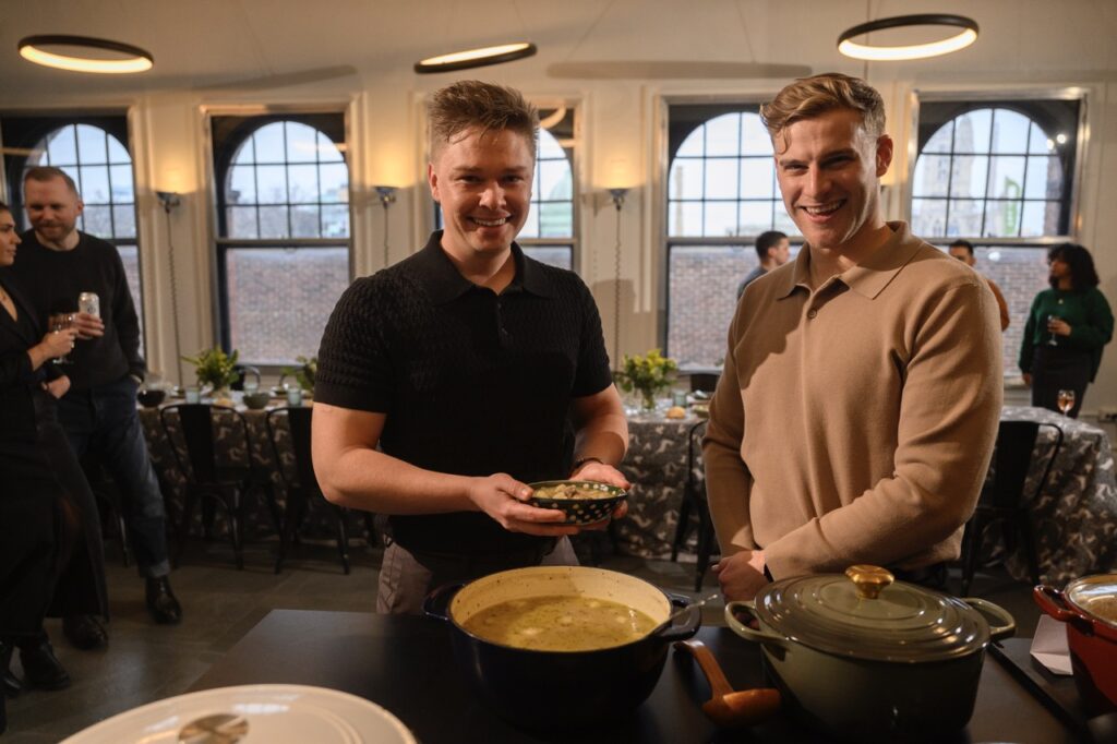 Two men gather around a pot of soup, bowls in hand.