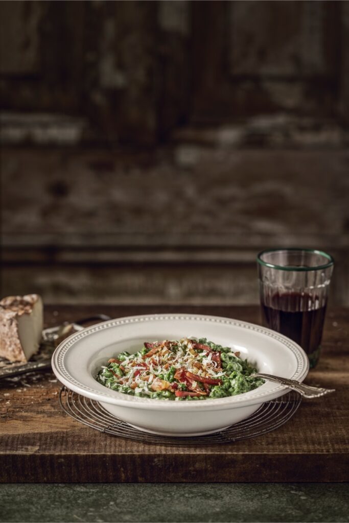 A deep white bowl of green spinach spätzle topped with crispy speck and grated cheese, next to a glass of red wine and a piece of cheese on a rustic wooden table.