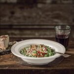 A deep white bowl of green spinach spätzle topped with crispy speck and grated cheese, next to a glass of red wine and a piece of cheese on a rustic wooden table.