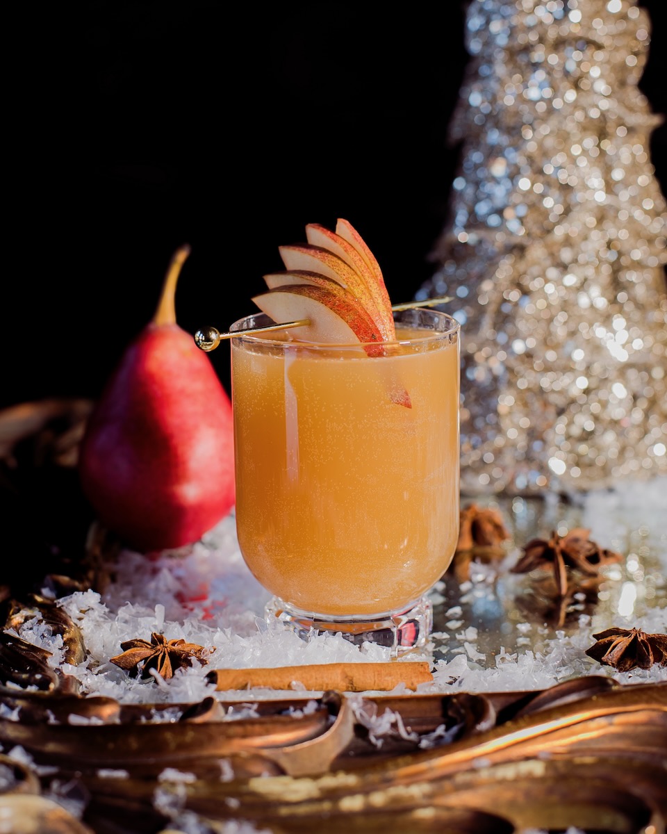 An orange Christmas mocktail staged with Christmas decorations and a red pear