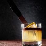 A cocktail for benoit blanc in knives out with a in a glass in a pale yellow color with a red float on top, lemon twist and knife through it all.