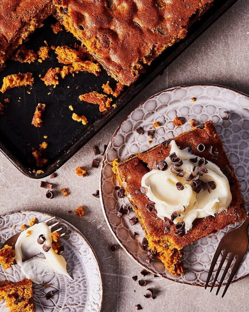 A moist pumpkin blondies topped with thick cream cheese frosting and chocolate curls, served on rustic plates next to the baking pan.