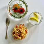 A celery root and grated carrot salad dressed with Vadouvan dressing, served on a clear plate with a fork, next to two glasses of water, one containing lemon slices and one with floating orange flowers and thyme.