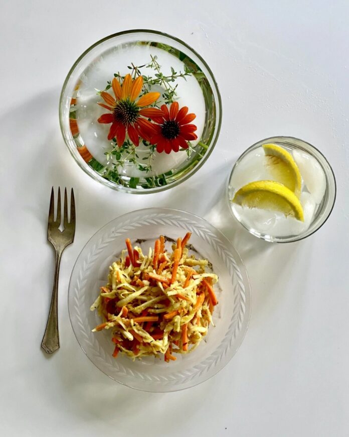 A celery root and grated carrot salad dressed with Vadouvan dressing, served on a clear plate with a fork, next to two glasses of water, one containing lemon slices and one with floating orange flowers and thyme.