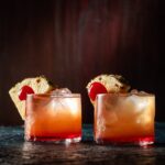 Two glasses of an orange cocktail with cherry and pineapple wedge garnishes.