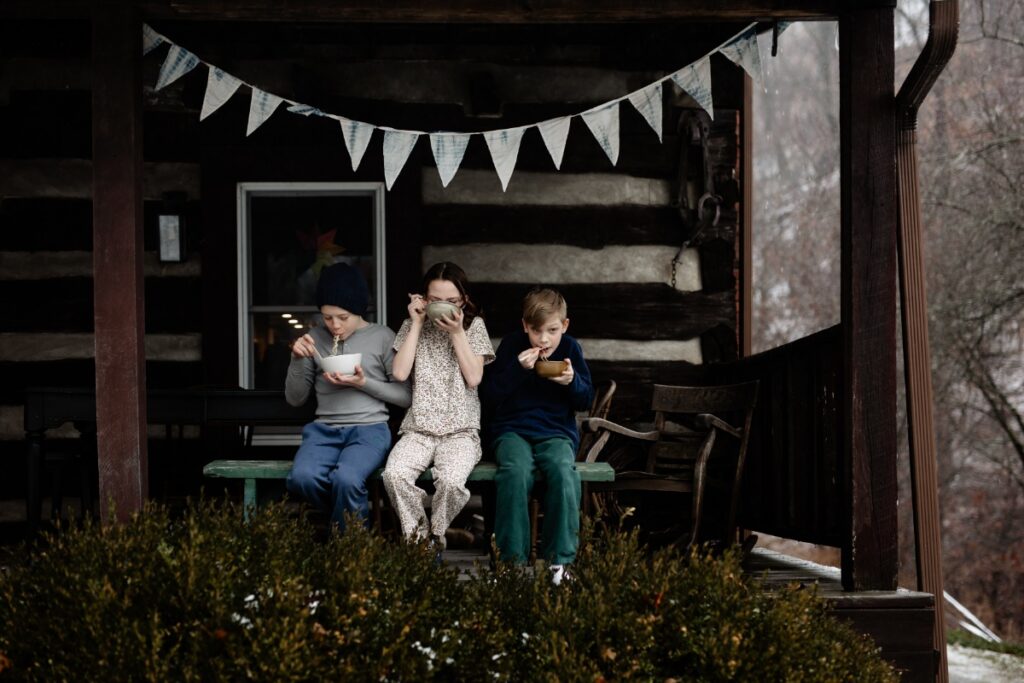 Three children sit on a rustic wooden porch bench, eating out of small bowls, with a white pennant banner hanging above them on the log cabin wall.