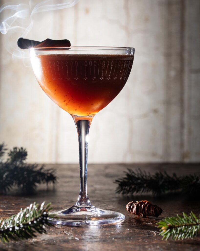 A coupe glass with a brown Three Wisest Men cocktail and a toasted cinnamon stick garnish as sprigs of pine trees lay on the table.
