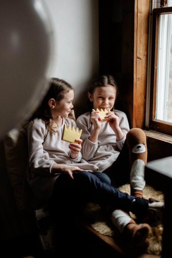 Two girls sit on a window seat, one biting a yellow Max crown cookie while the other holds one, wearing long-sleeved shirts and cozy socks, by a bright window.