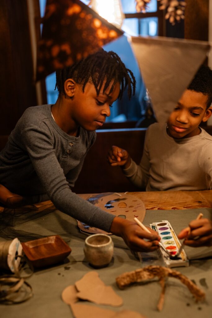 Two young boys are seen close up making "Wild Thing" masks, one reaching to paint with a watercolor palette and the other looking on, in dim light.