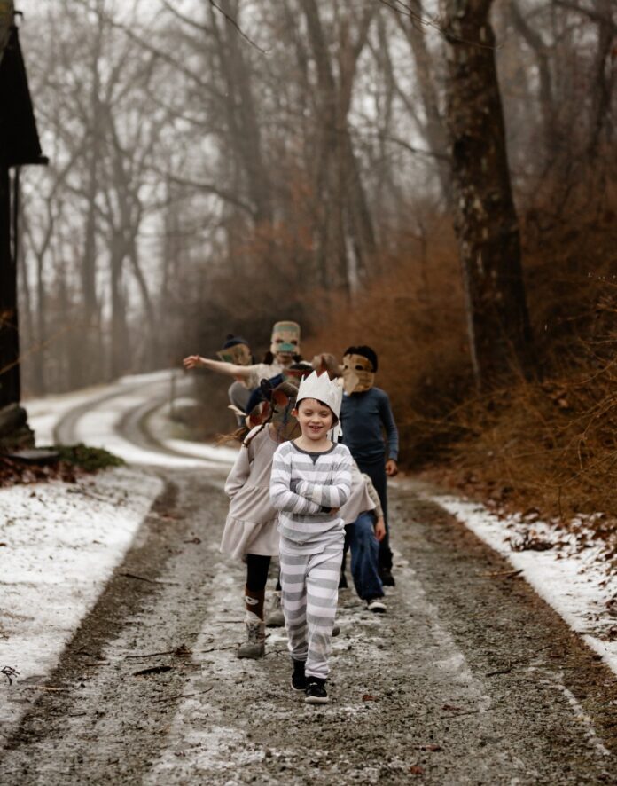 A line of young children dressed in pajamas and animal masks walking down a snowy, muddy path in the woods, with the boy in front wearing a crown and striped pajamas.