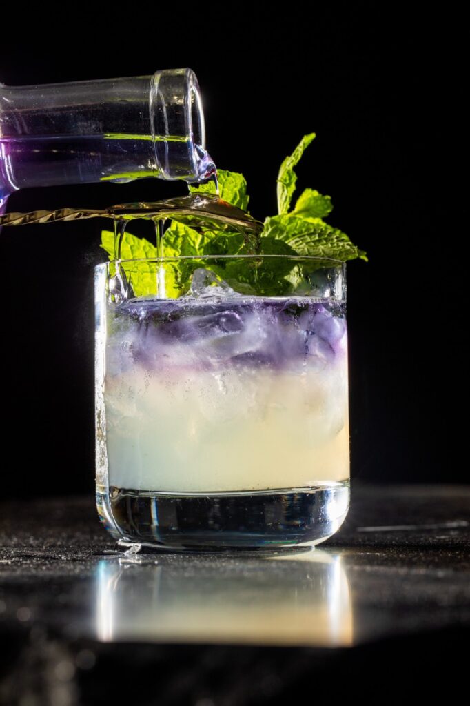 A small shot of pueple gin pours into a Wicked: For Good cocktail with a foggy yellow base and mint leaves on top.