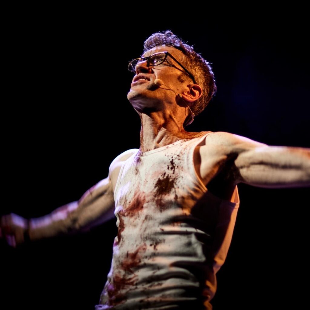 An actor on stage in a white tank top with blood. 