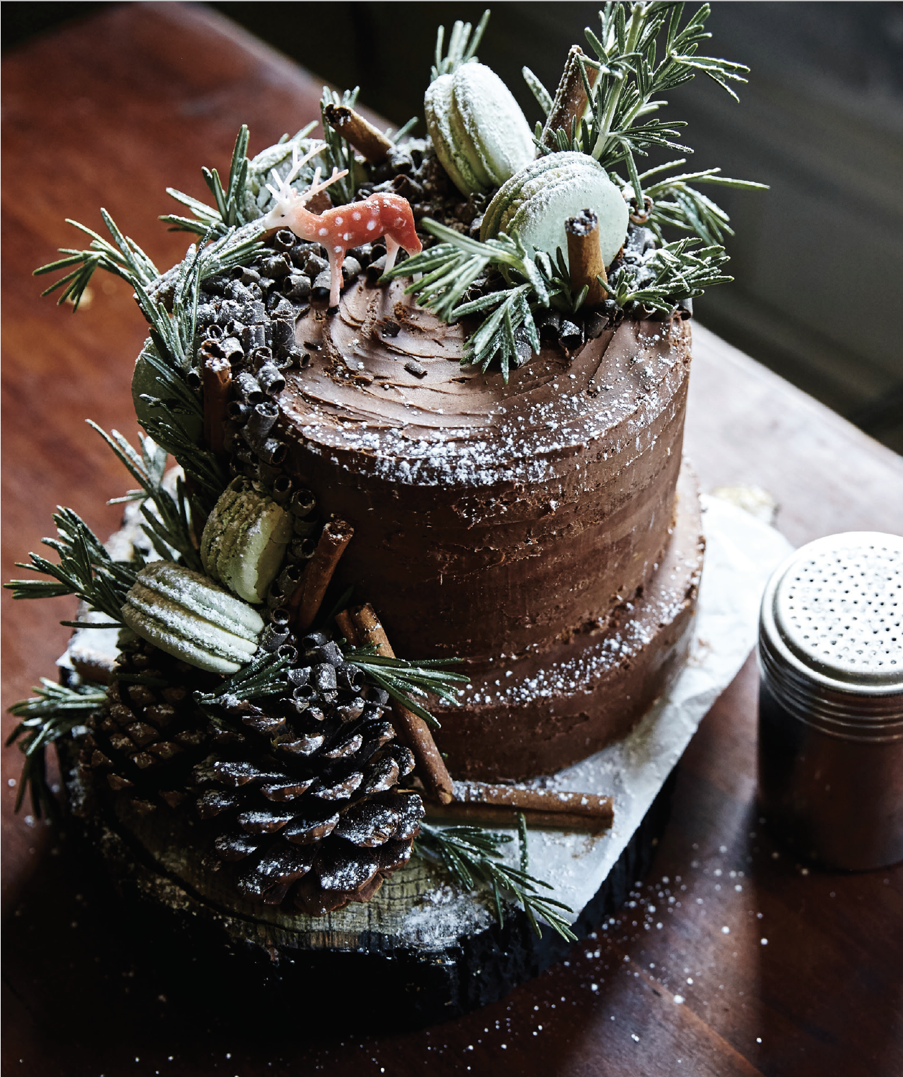 An Whole Grain Apple Cider Layer Cake covered in chocolate frosting and white decor like pinecone and cinnamon sticks.