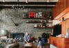 A Mid-Century Retreat in Upper Lawrenceville A mid-century modern living room with a long, curved gray sofa facing a stacked stone fireplace, featuring a low wooden coffee table, wood paneling, wall-mounted shelving with decor, and a wooden floor with a gray shaggy rug.