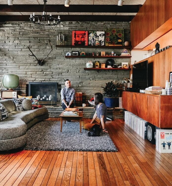 A mid-century modern living room with a long, curved gray sofa facing a stacked stone fireplace, featuring a low wooden coffee table, wood paneling, wall-mounted shelving with decor, and a wooden floor with a gray shaggy rug.