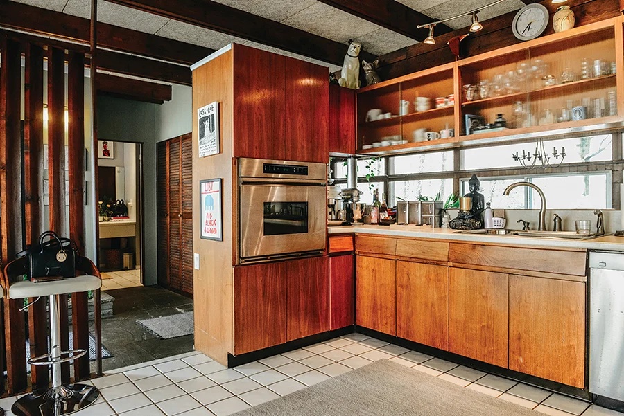 A mid-century modern kitchen with warm wood cabinets, white tile floor, stainless steel appliances, a barstool with a purse, and a pass-through opening on the left, demonstrating the home's original 1957 design.