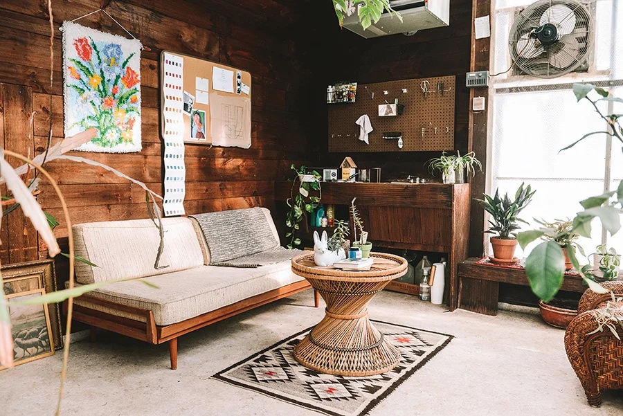 A mid-century modern living room area featuring a gray, two-cushion sofa with plaid and black pillows, a collection of electric and acoustic guitars mounted on a paneled accent wall, and a floor lamp.
