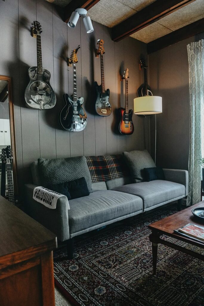 A living room with six guitars hanging on a wood-paneled wall above a light gray couch with dark pillows and a plaid throw blanket, showcasing a music-themed interior design.