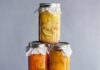 How to Make a Shrub at Home A vertical stack of six glass mason jars filled with cold-processed fruit and vegetable shrub mixtures for cocktails or drinks, including yellow fruit, orange puree, dark berries, and light colored chunks, each topped with white cloth covers.