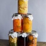 A vertical stack of six glass mason jars filled with cold-processed fruit and vegetable shrub mixtures for cocktails or drinks, including yellow fruit, orange puree, dark berries, and light colored chunks, each topped with white cloth covers.