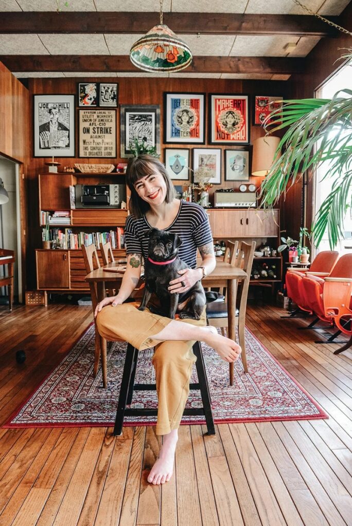 A smiling woman, Jane Hallinan, sitting on a high stool holding a black pug in a mid-century modern living and dining room with hardwood floors, wood-paneled walls, a colorful gallery wall of framed art, and modern wooden furniture.