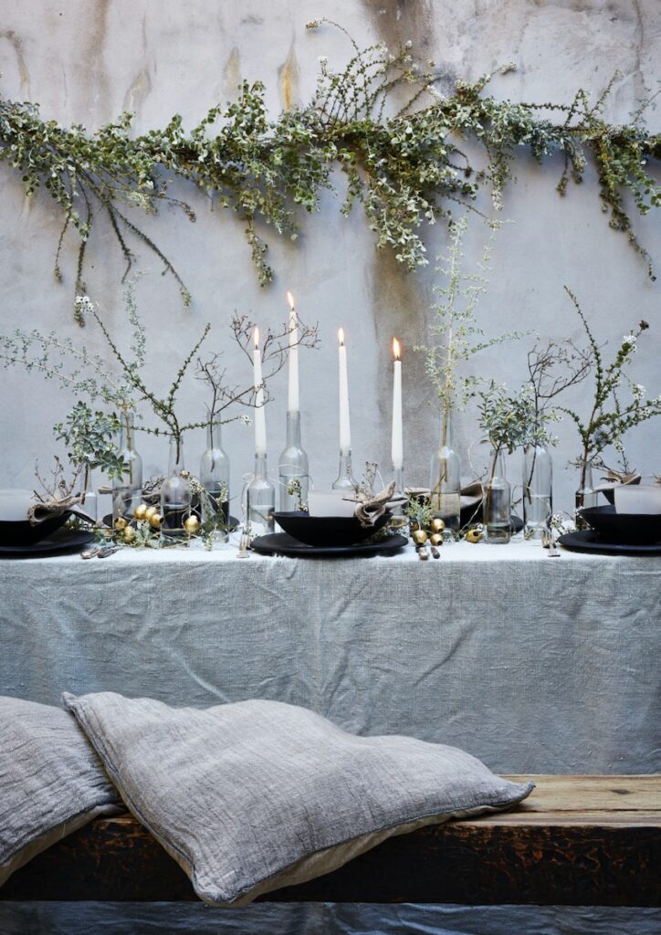 A table set with candles and sprigs of Christmas plants, surrounded by grey throw pillows