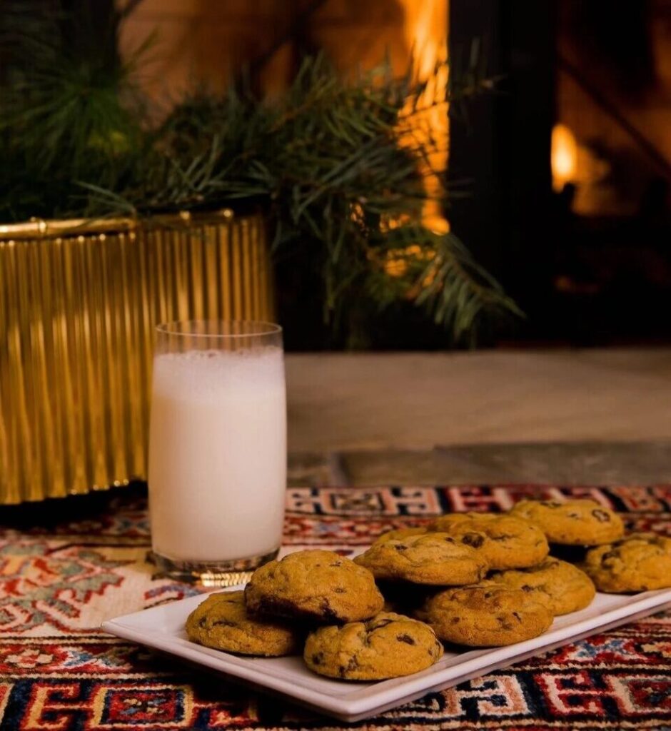 A plate of warm chocolate chip cookies and a glass of milk on a festive rug, with pine branches and a fireplace glowing in the background.