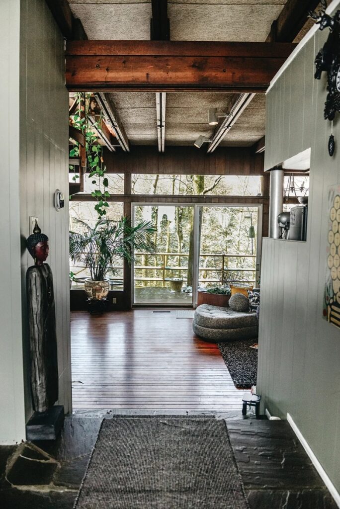 The entryway into a mid-century modern home with dark hardwood floors, wooden ceiling beams, and a wall of windows offering a forested view from the deck, featuring a round lounge seat and a large potted plant.