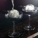 Water pours over an ice ball in a martini glass.
