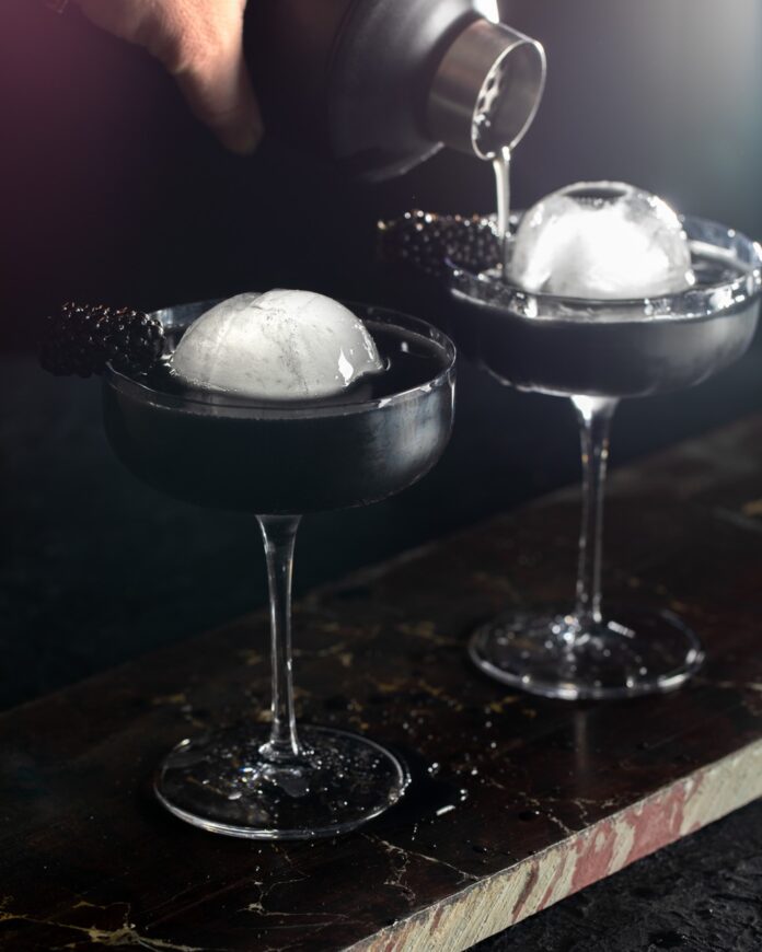 Fallout Season 2 Cocktail TABLE Magazine 2 Two martini glasses with two sphere ice balls as a person pours something into the back one.