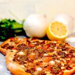 Two round flatbreads topped with spiced ground meat, diced tomatoes, and pine nuts (likely lahmajoun or sfiha), placed on a gray surface next to white onions, fresh parsley, and a cut lemon.