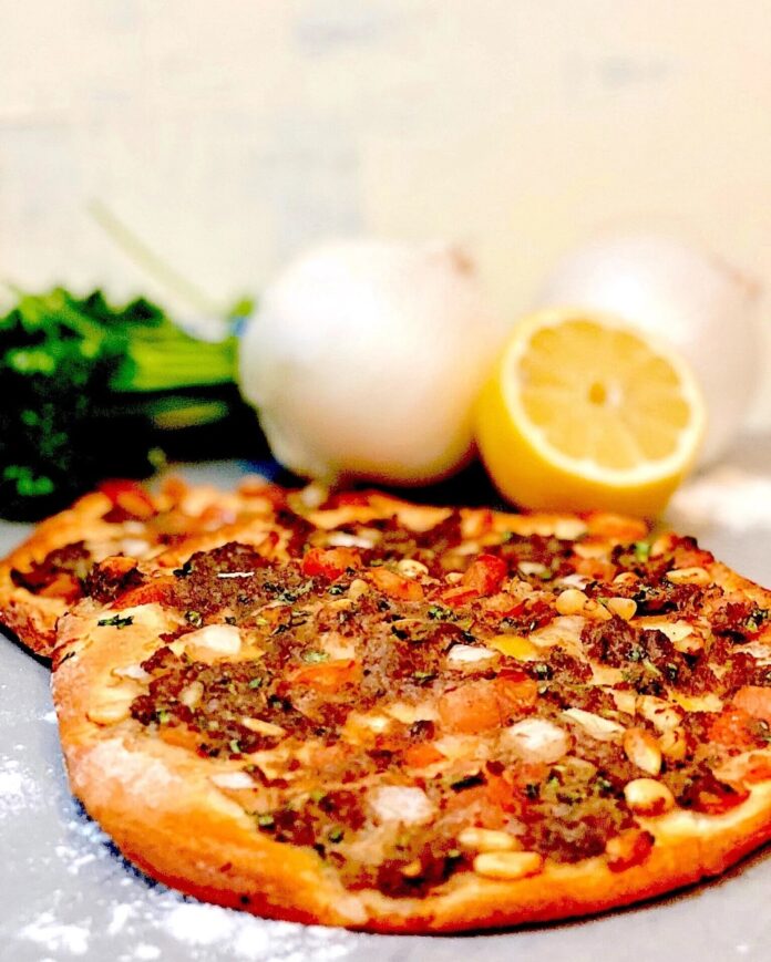 Two round flatbreads topped with spiced ground meat, diced tomatoes, and pine nuts (likely lahmajoun or sfiha), placed on a gray surface next to white onions, fresh parsley, and a cut lemon.