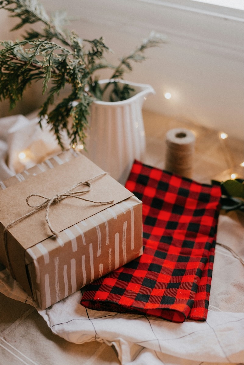 A brown wrapped gift sits on a white cloth table with a plaid hanker-chief beneath it and a pine branch in a ceramic pitcher.