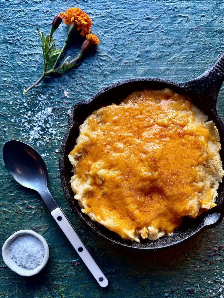 A cast iron pan of Green Chile and Cheddar Mashed Potatoes with a spoon, salt, and orange flowers beside the pan.
