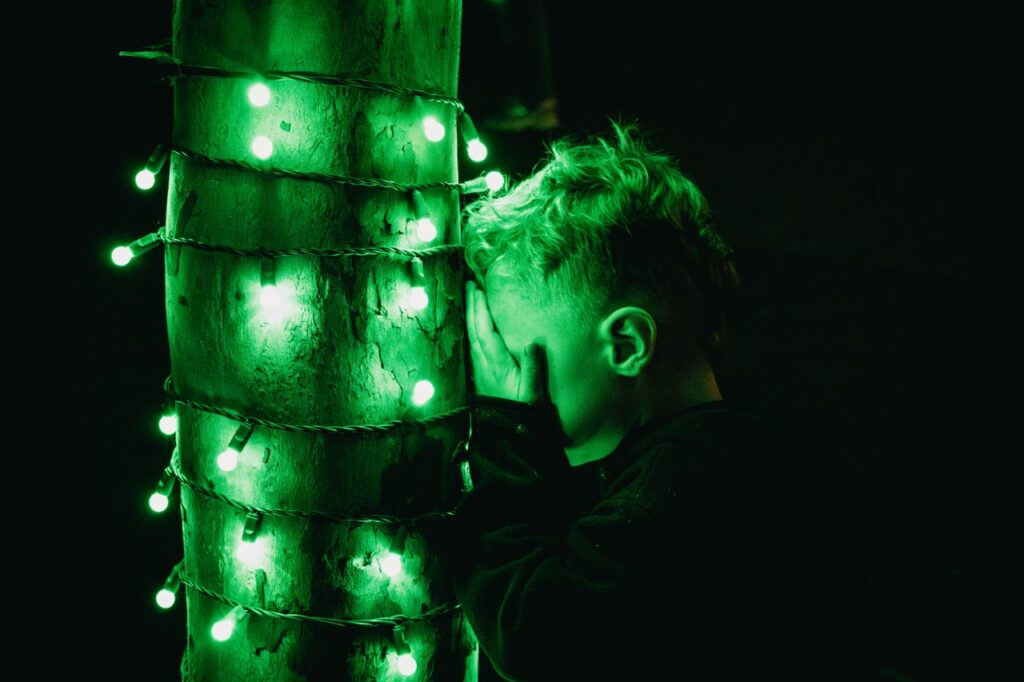 A young boys covers his eyes and sets his head against a green lit tree.