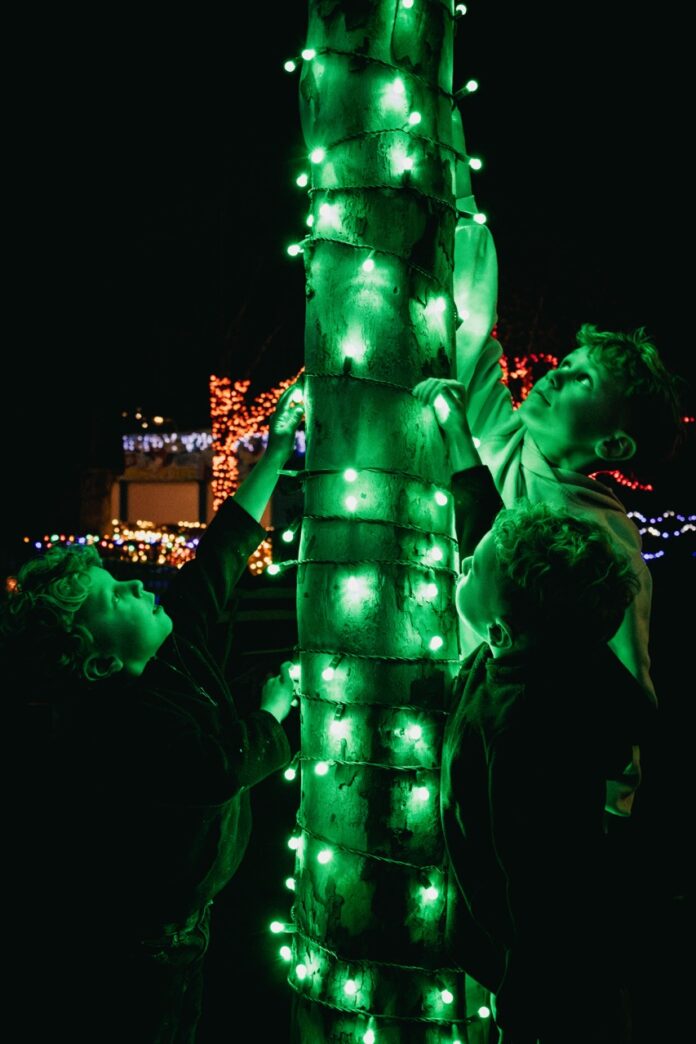 Three boys play with a tree wrapped in green lights.