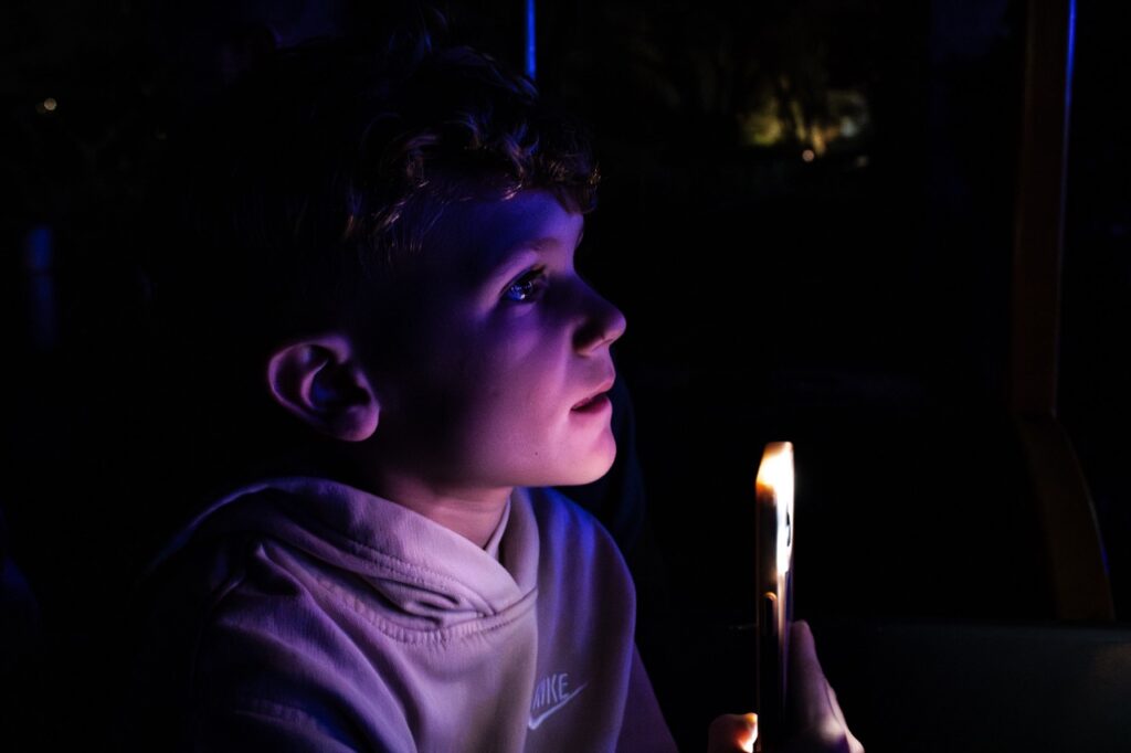 A young boy looks as his face is lit by his phone flashlight.