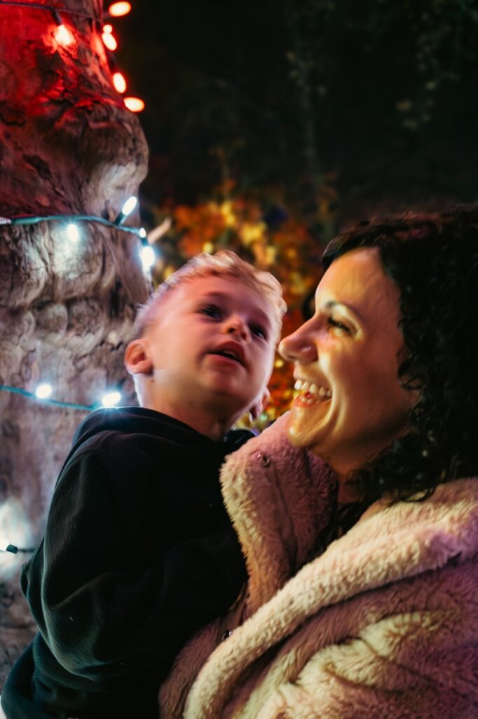 A mom holds a young boy up to a lit tree.